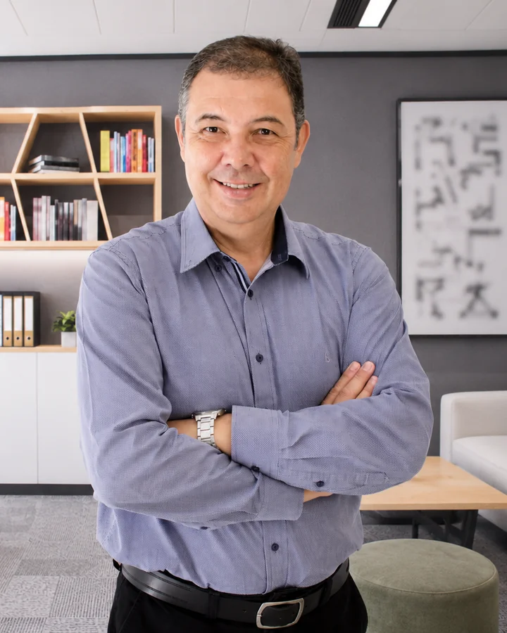 Eng. Pedro Rodrigues Neto, sócio-diretor da Rodrigues Neto, sorrindo de braços cruzados, de camisa azul, em um escritório com estante e sofá ao fundo